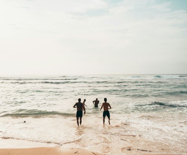 Three people running into the ocean from a sandy beach under a partly cloudy sky.