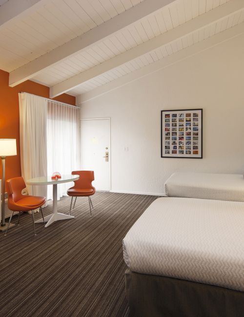 A modern hotel room with two beds, a flat-screen TV, a small table, and chairs. Orange accent wall adds warmth to the decor.