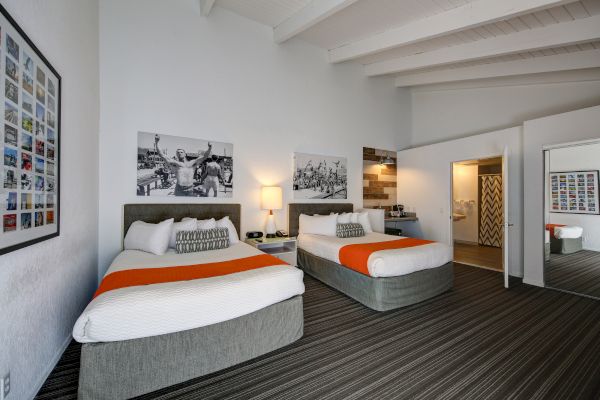The image shows a hotel room with two beds, orange accents, framed wall art, a nightstand with a lamp, and a view into a bathroom.