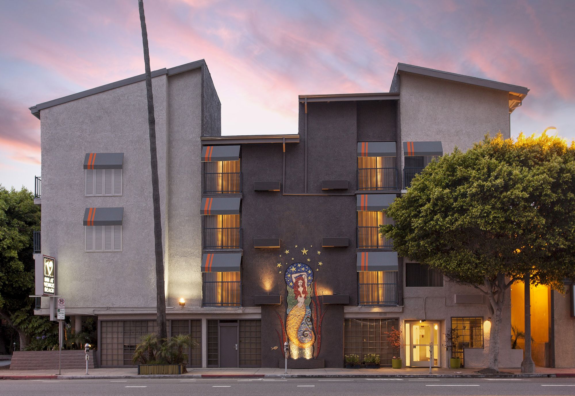A modern building with colorful mural art and large windows, under a sunset sky, surrounded by trees and a street.