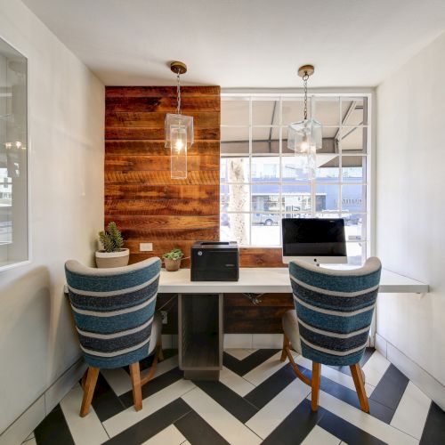 The image shows a modern workspace with two chairs, a computer, a printer, and plants on a white desk. There's a window and striped floor.