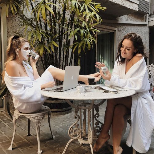 Two people in robes relax at a patio table with coffee, a laptop, and beauty products, enjoying a casual pampering session.