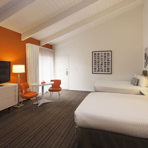 The image shows a modern hotel room with two beds, an orange accent wall, a TV, a small table with orange chairs, and decorative wall art.