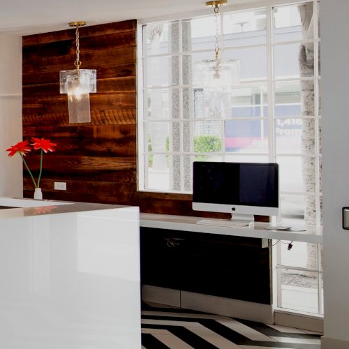 The image shows a modern office space with a white desk, computer, red flowers, wood paneling, pendant lights, and a large window.