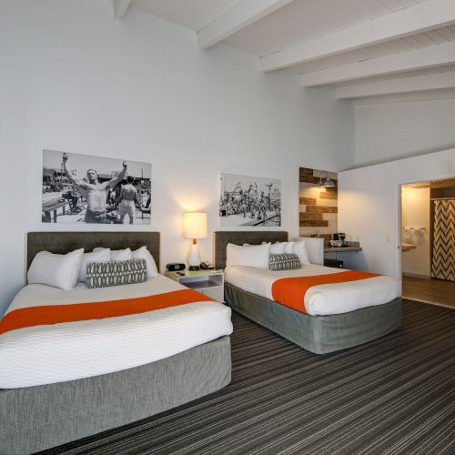 This image shows a modern hotel room with two beds, orange accents, framed art, and an adjoining bathroom.