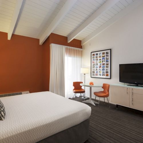 Bright hotel room with a large bed, lamp, TV, orange chairs, and a table under white beams and artwork on the wall.
