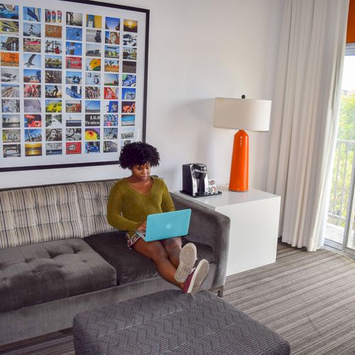 A person is sitting on a sofa with a laptop in a modern room, featuring bright decor, a gallery wall, and a window view.