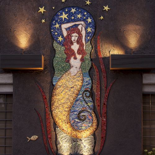 A mural of a mermaid with a red tail set against a starry night sky, illuminated by lights, and adorned with ocean-themed details.