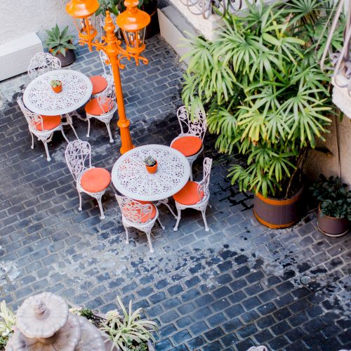 A charming patio with white ornamental tables and chairs adorned with orange cushions, surrounded by potted plants and an orange lamp post.