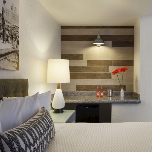 A cozy hotel room with a bed, decorative pillows, a lamp, vintage photo, counter with flowers, drinks, and a textured wood wall accent.